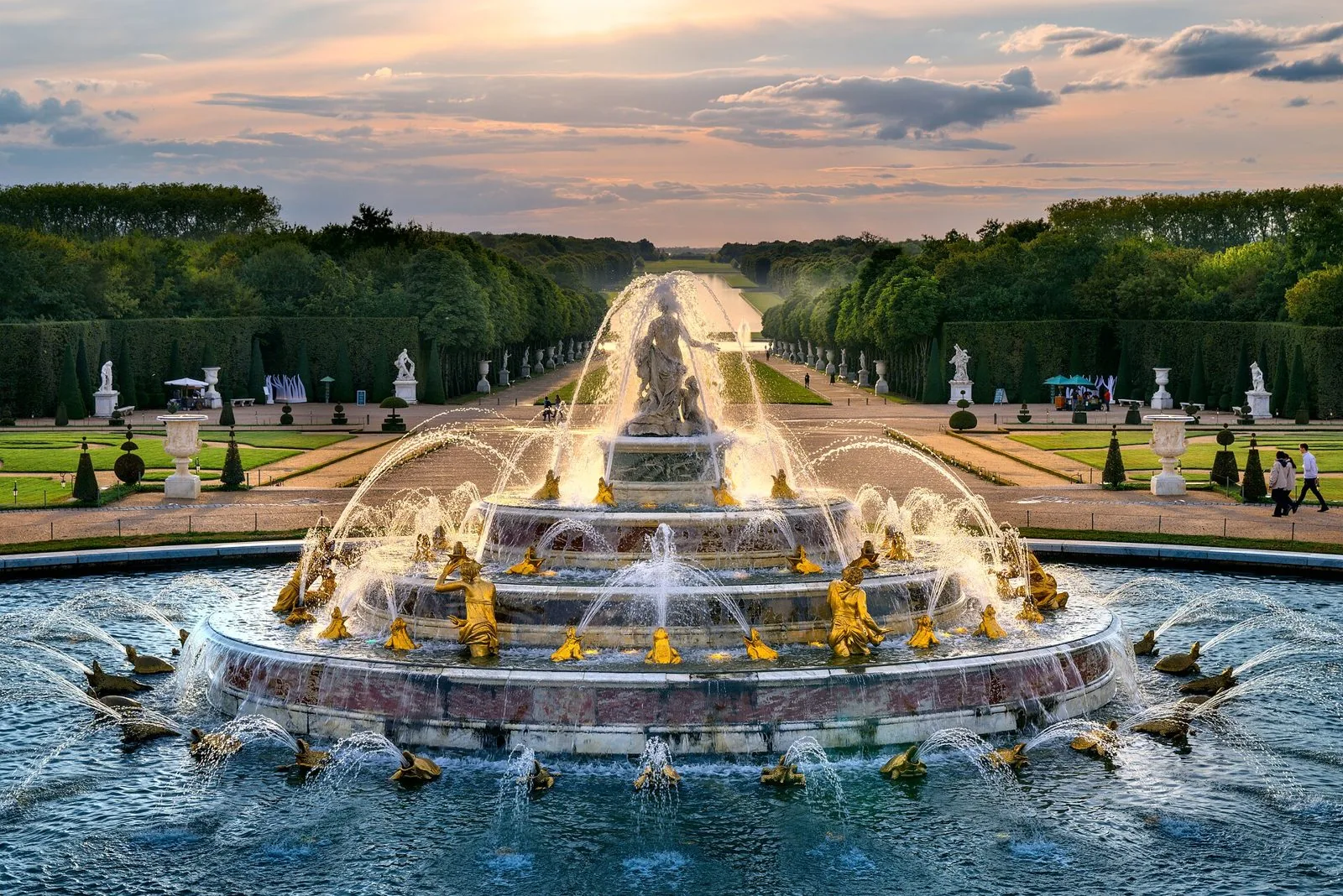 La fontana di Latona nei giardini di Versailles con i getti d'acqua in funzione e le statue dorate, sullo sfondo il Tapis Vert e il Grand Canal al tramonto